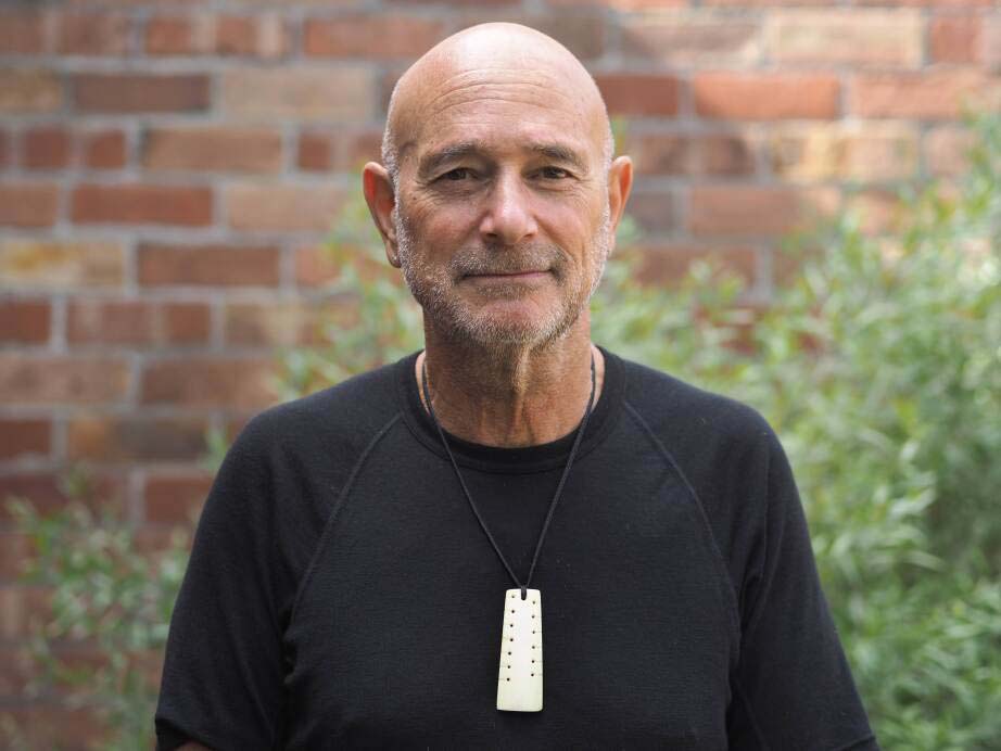 A man stands in front of a brick wall and a bush.