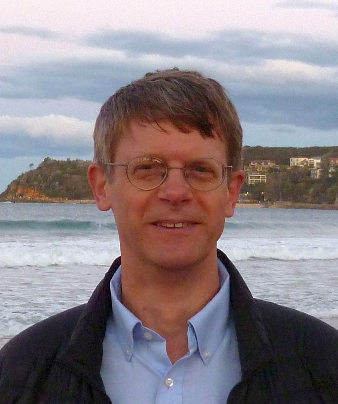 Chet Van Duzer in a blue shirt and jacket standing in front of the ocean