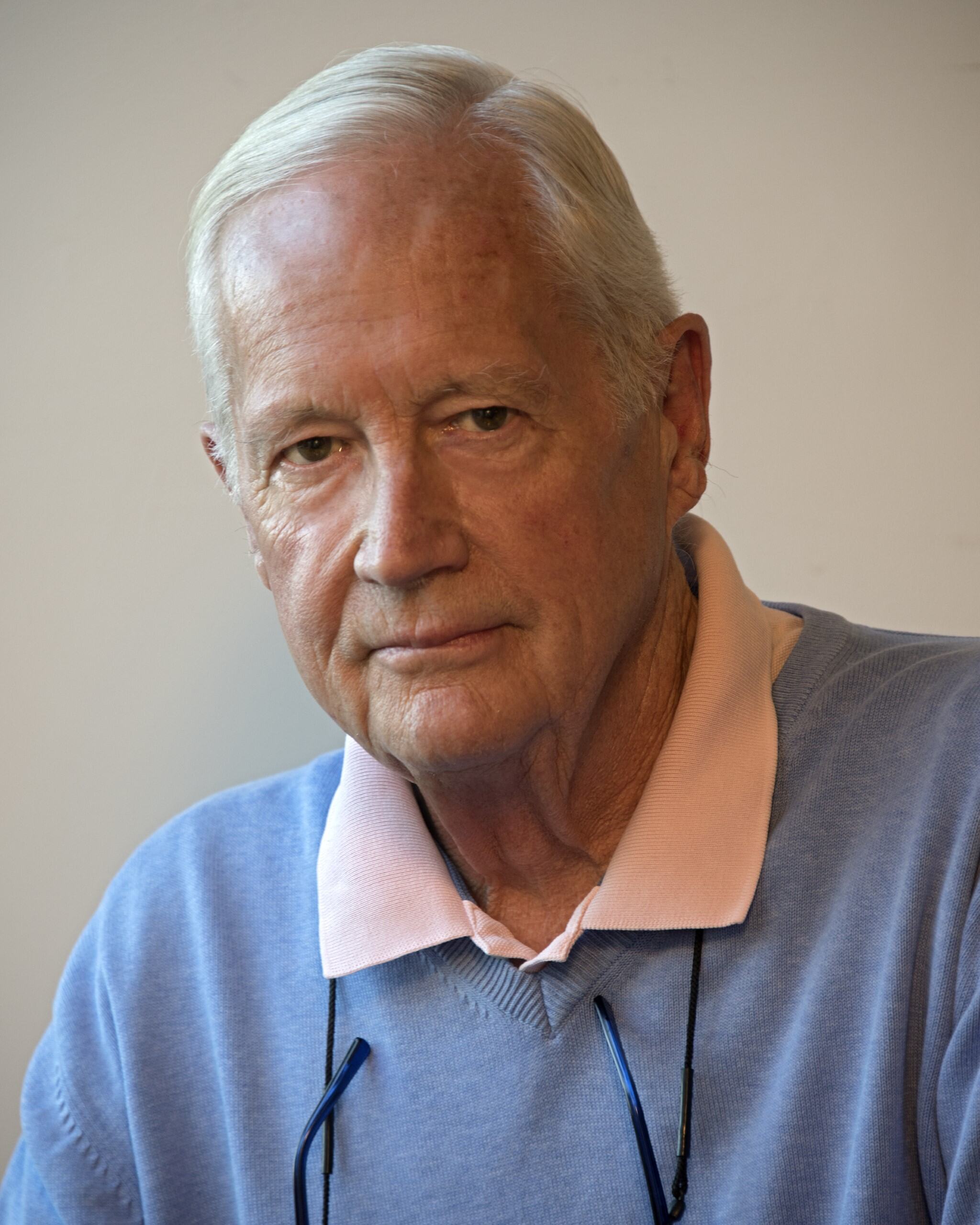 A portrait of Robert Macklin wearing a sky-blue sweater, with the pink collar of the undershirt visible over the swetaer's collar. 