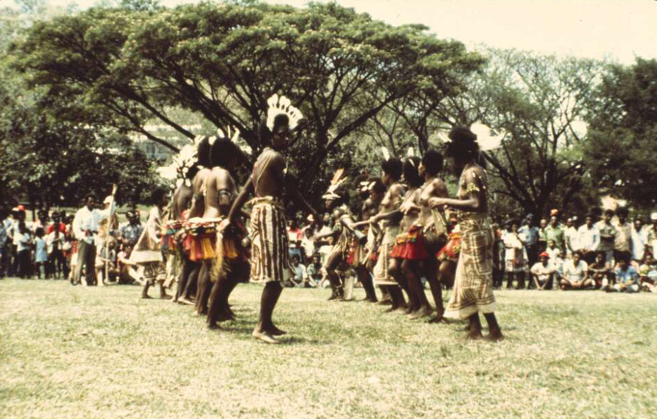 Papua New Guinean dancers wearing traditional dress and performing for a crowd of onlookers, in an outdoor setting