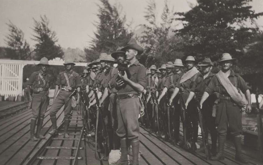 Australian Naval and Military personnel standing in two rows. The man at the end of the front row is holding a little black dog.