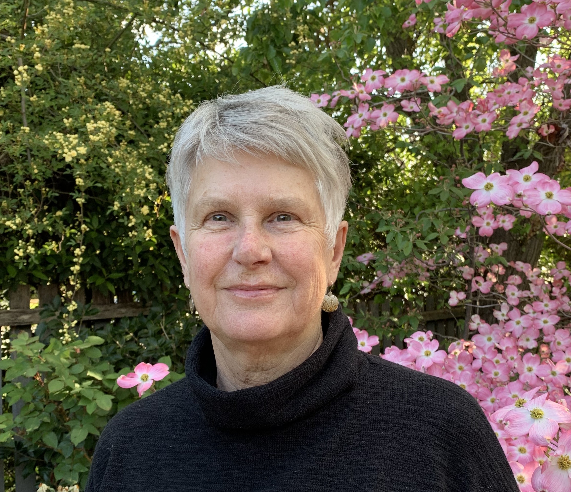 Helen Ennis wearing a black turtleneck sweater, standing in front of a pink flowering rhododendron