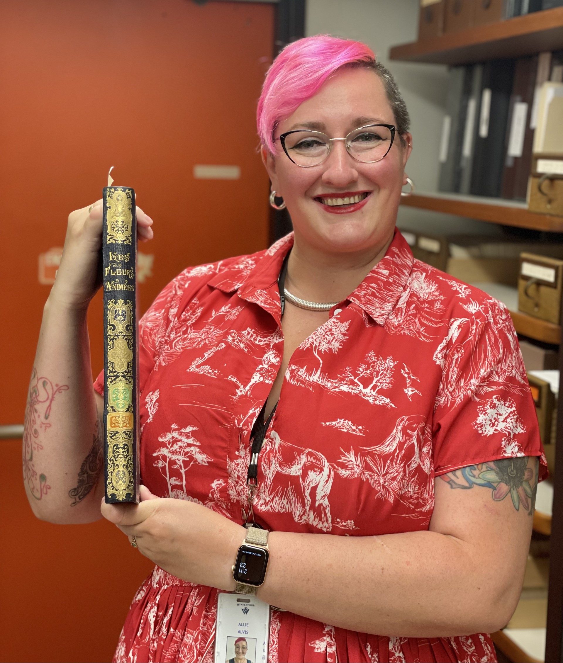 A pink haired person with glasses wearing a red dress holds up an old book