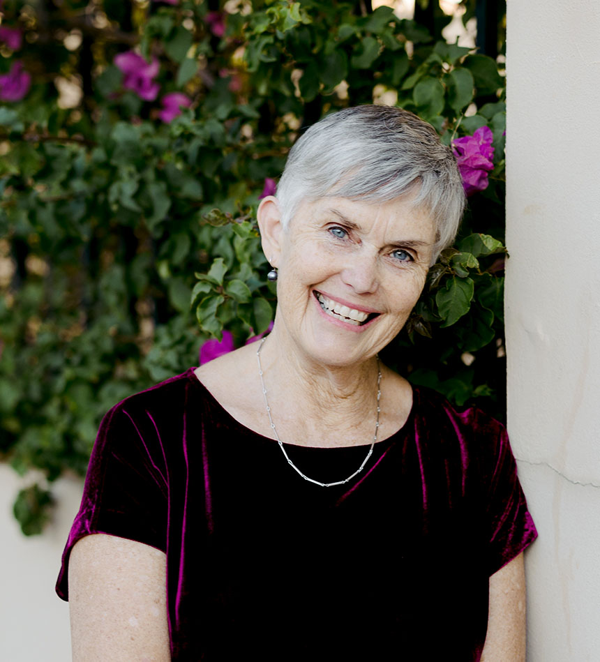 A portrait of Dianne Wolfer. She wears a burgundy dress and a silver chain, and stands infront of a rosebush garden