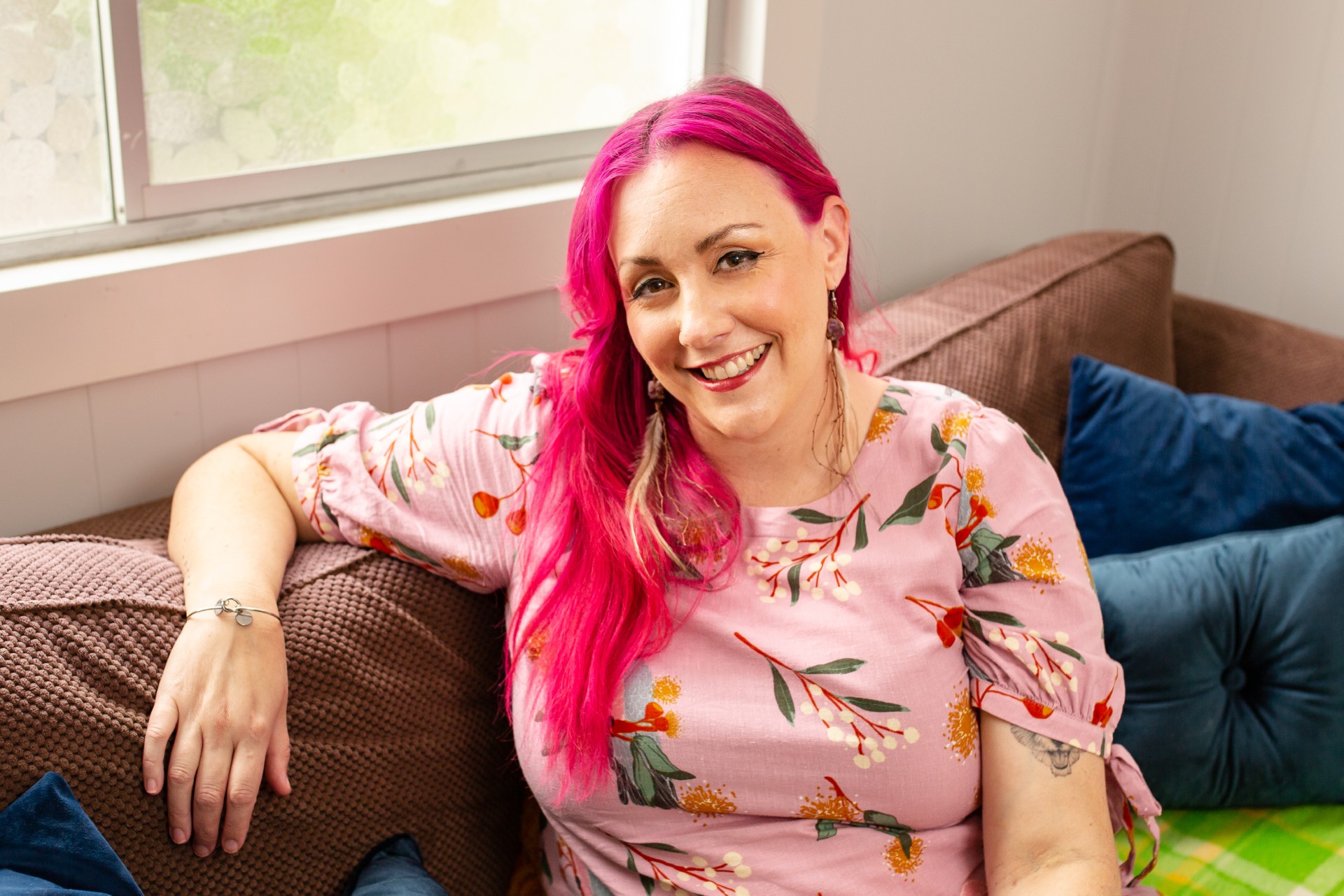 Woman with bright pink hair, wearing a pastel pink shirt, sitting on a couch by a window
