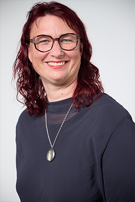 A portrait of a smiling woman wearing glasses, a dark blue shirt and a silver pendant necklace
