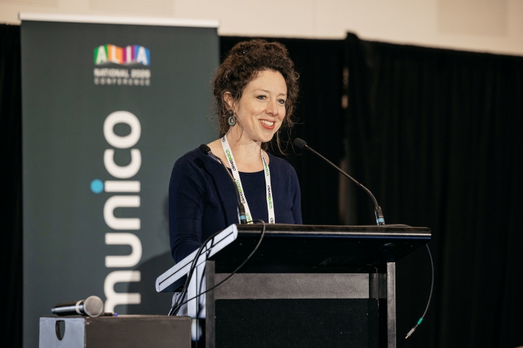 A smiling woman with curly brown hair is giving a presentation at a conference, standing behind a podium.