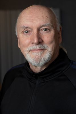 A man with a white beard, looking directly at the camera. He is wearing a black jacket.