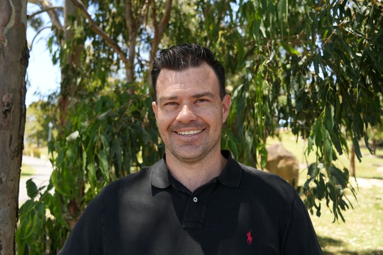A smiling man with dark hair is pictured outdoors in a park or wooded area. He is wearing a black polo shirt with a small red logo visible on the chest. The background is filled with lush green trees and bright daylight.