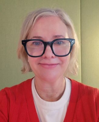 A smiling woman with blonde hair and black-rimmed glasses, wearing a red cardigan over a white top.