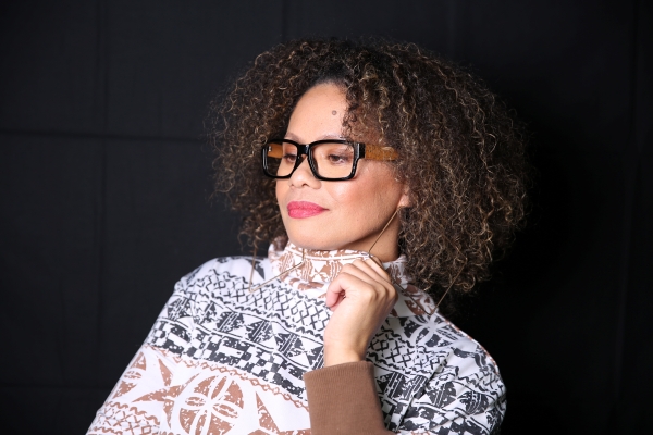 A woman with voluminous curly brown hair and glasses, wearing a patterned white and black top. She is looking slightly to the side with a thoughtful expression against a dark background.