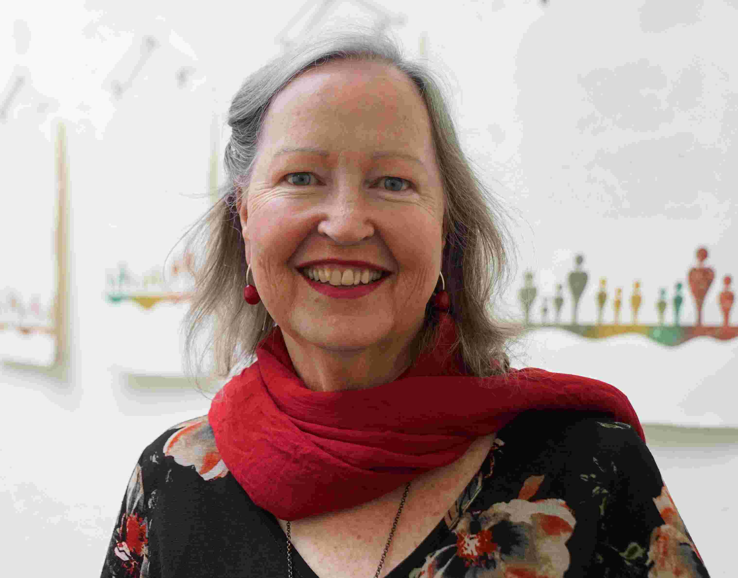 A smiling woman wearing a red scarf and red lipstick, pictured against a blurred background of artworks hung on a gallery wall