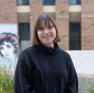 A young woman with straight brown hair and bangs wearing a black turtleneck sweater, standing outdoors in front of a modern brick building.