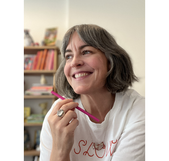 A smiling woman with short gray hair looks upward and to the side, holding a pink pencil. She wears a white t-shirt with red lettering and a necklace, with a bookshelf visible in the blurred background.