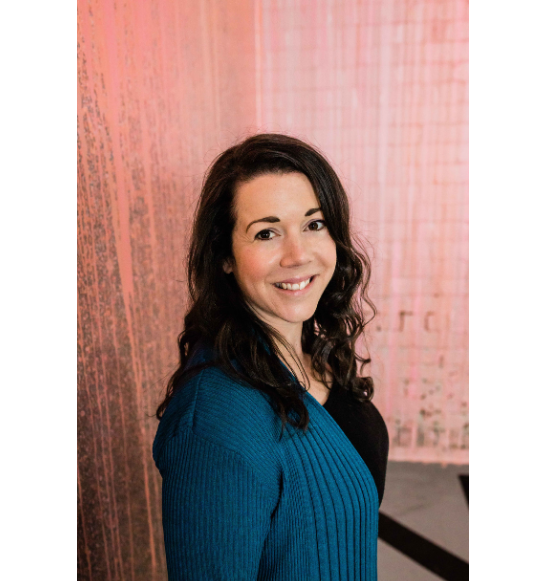 A smiling woman with dark, wavy hair wearing a blue ribbed cardigan over a black top, standing against a textured pink wall.