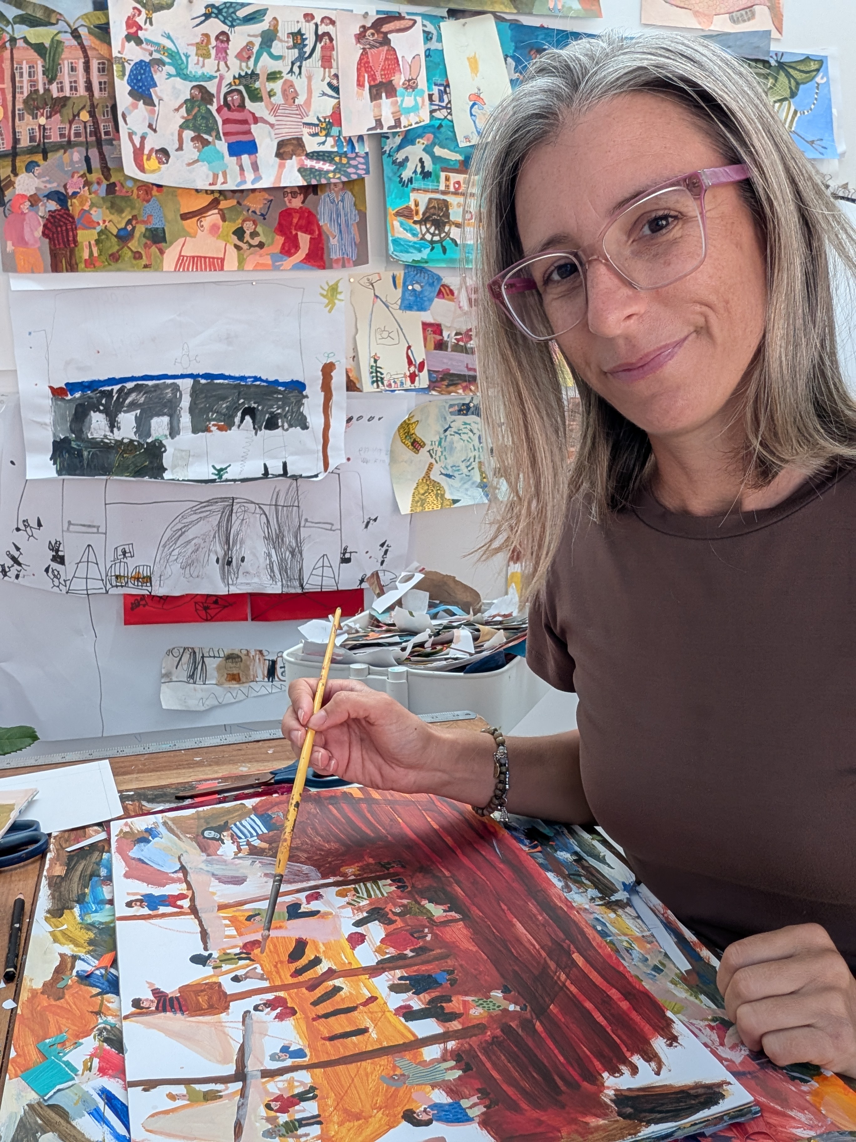 Person seated at an art desk painting a colourful illustration, with children’s artwork and sketches displayed on the wall behind.