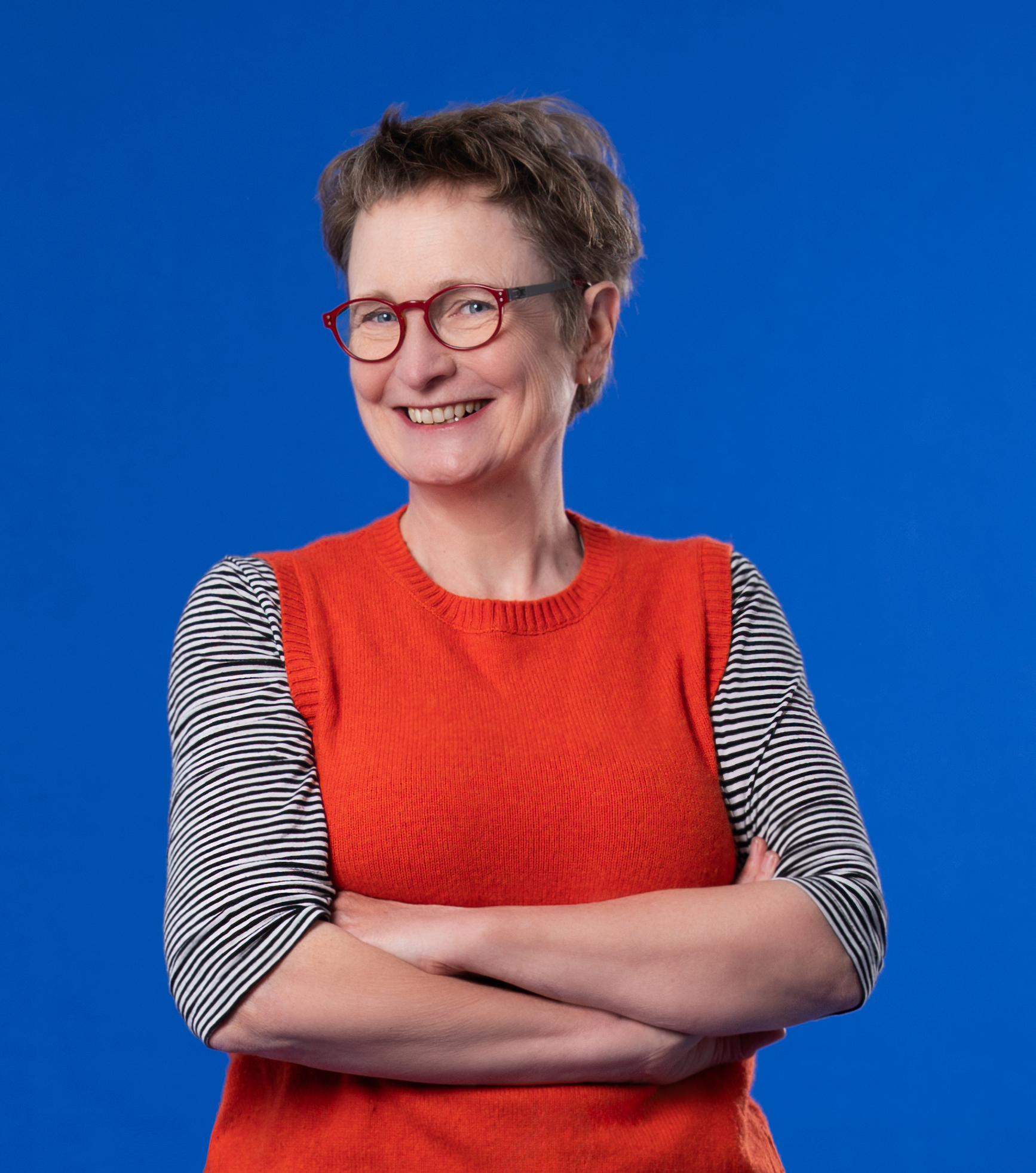A smiling woman with short dark hair wears red-framed glasses, a grey top and red vest and is pictured against a bright blue background