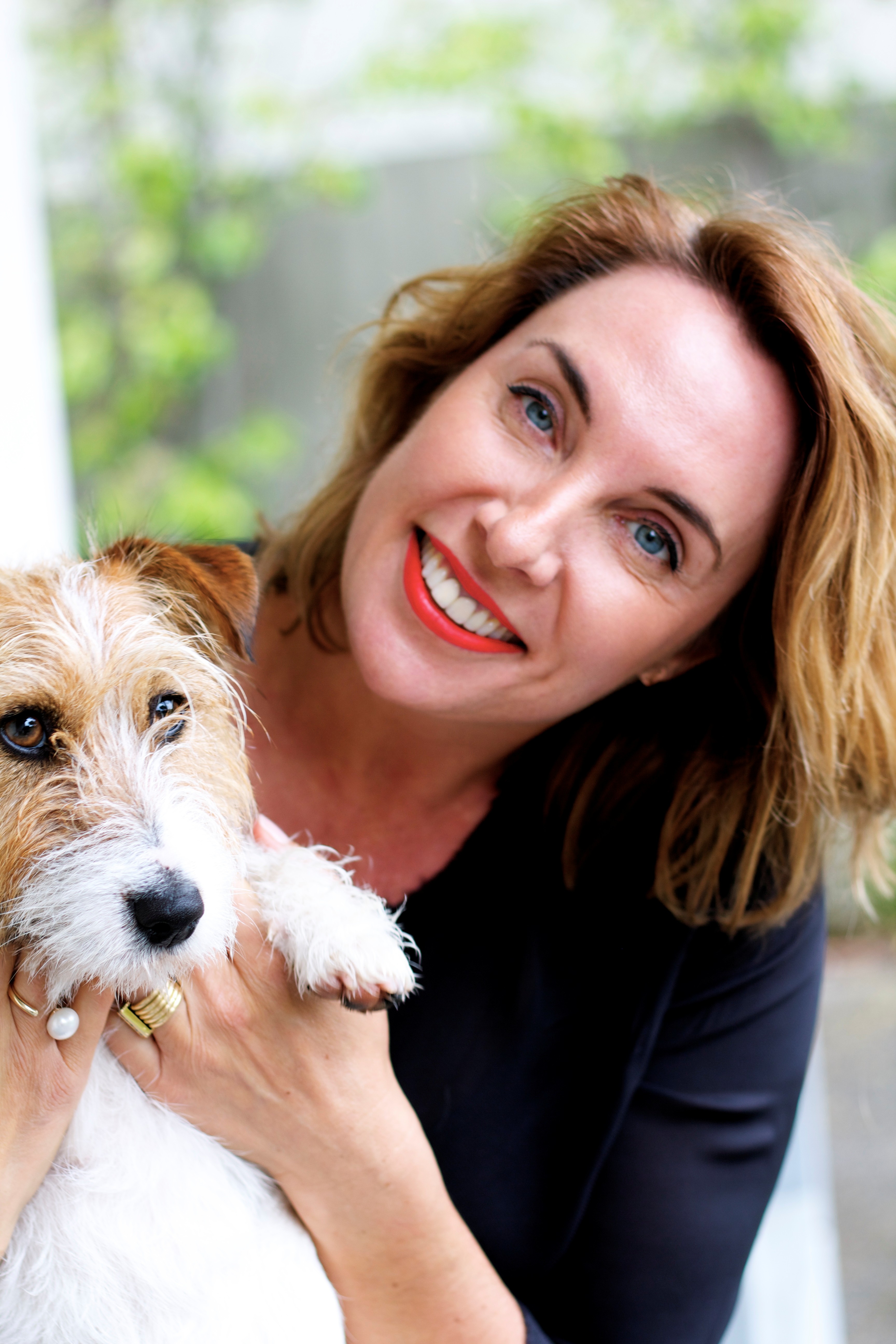 A smiling woman with red hair and bright red lipstick holds a small, scruffy terrier-like dog. She looks up at the camera with a joyful expression while cradling the dog, which has white and tan fur and dark eyes. The background is softly blurred greenery.