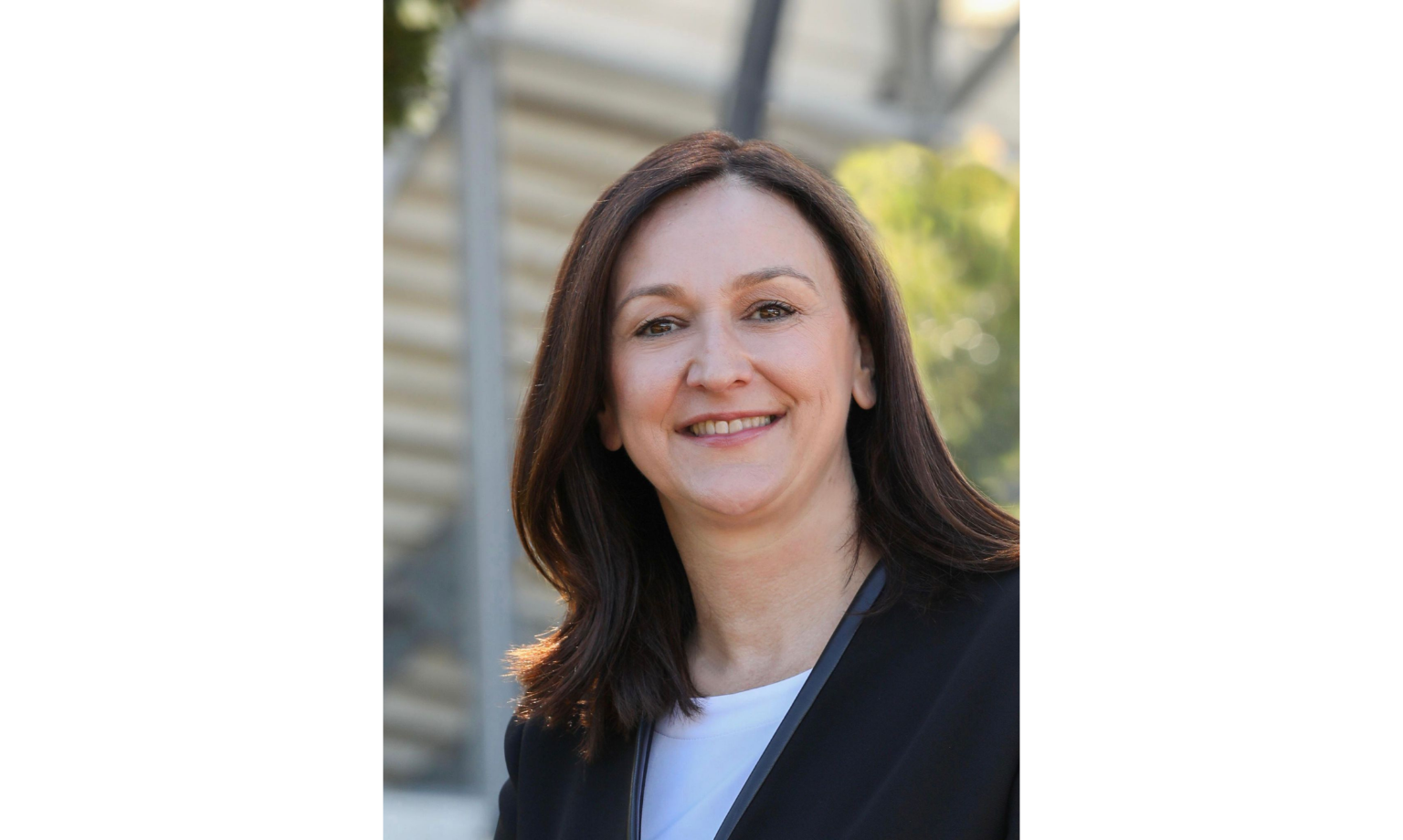 A smiling woman with shoulder-length brown hair wearing a black blazer over a white shirt, standing outdoors with a blurred background of greenery and architecture.