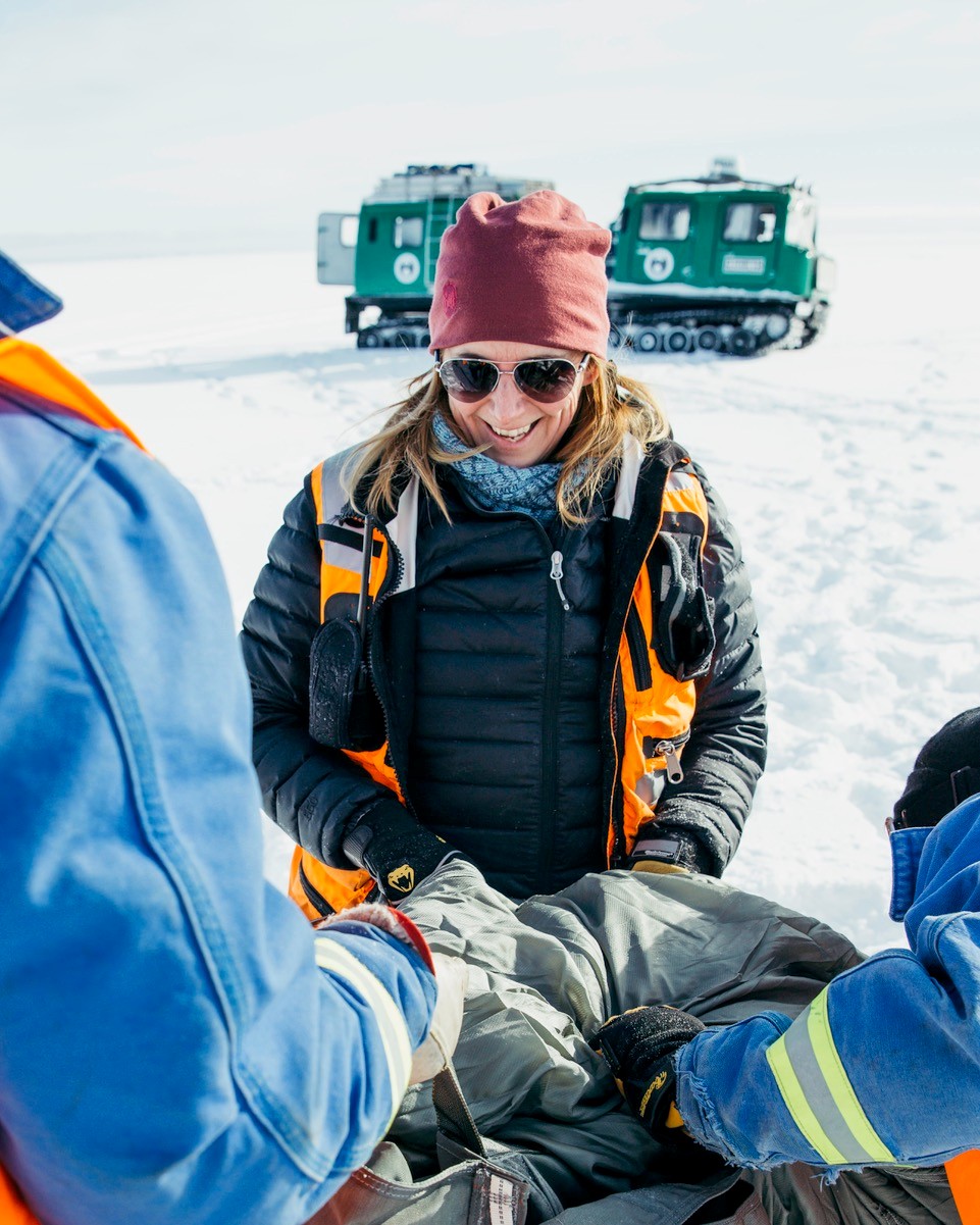 A smiling woman in winter gear, including a red beanie and sunglasses, is seated in the snow and appears to be interacting with others. She is wearing a black and orange insulated jacket and is surrounded by people in blue jackets with reflective stripes