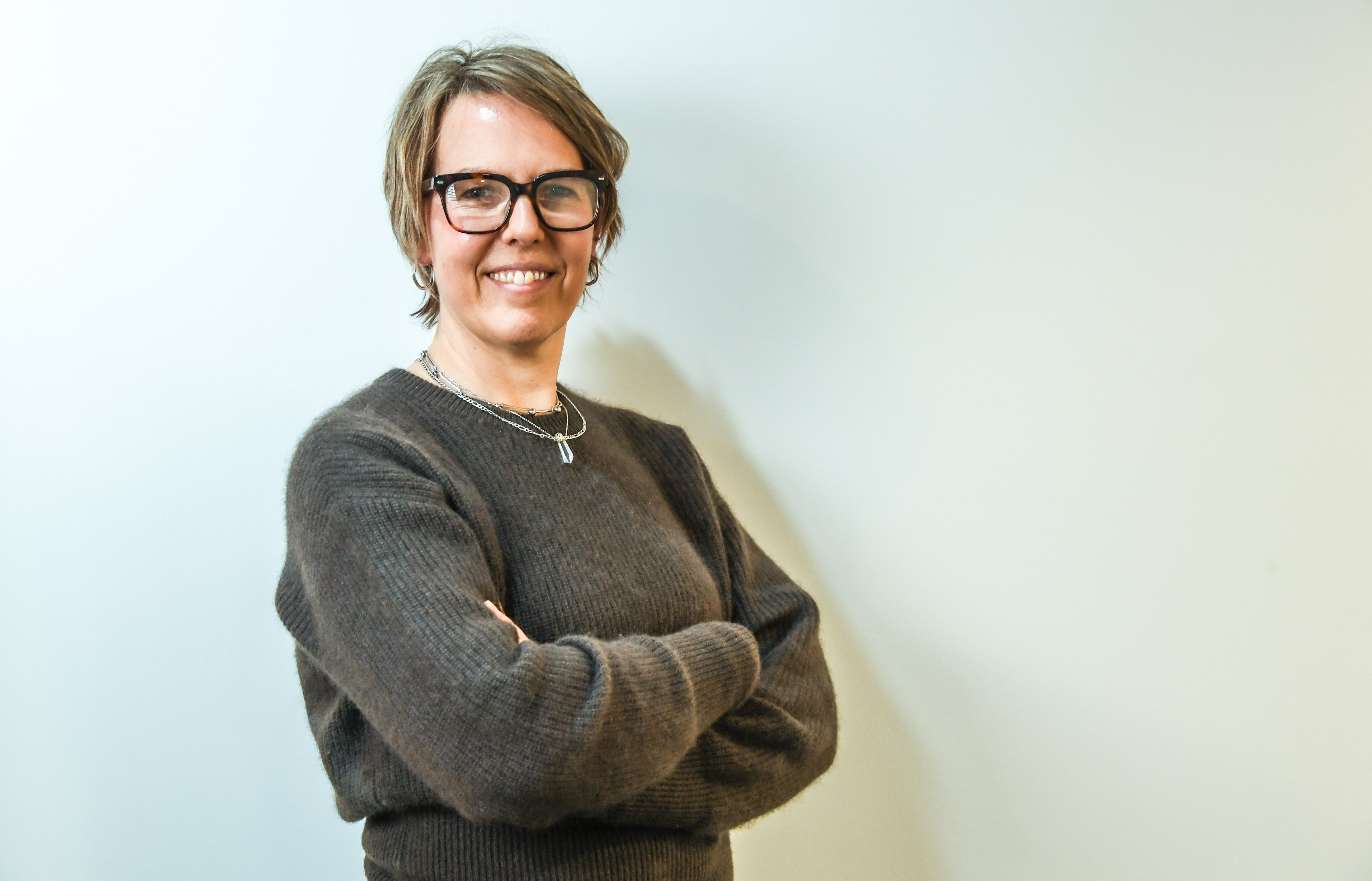 A smiling woman with short hair is wearing a grey jumper and glasses and is pictured in front of a white wall