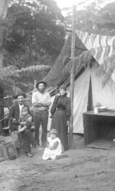 A black and white photograph of a family dressed in late 1800s style fashion. The are standing in a clearing in a forest and are standing in front of a tent.