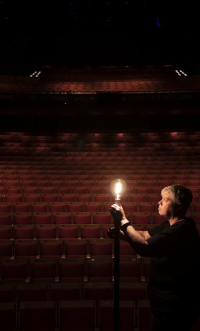 A person stands in an empty darkened theatre, illuminated by a single light bulb.