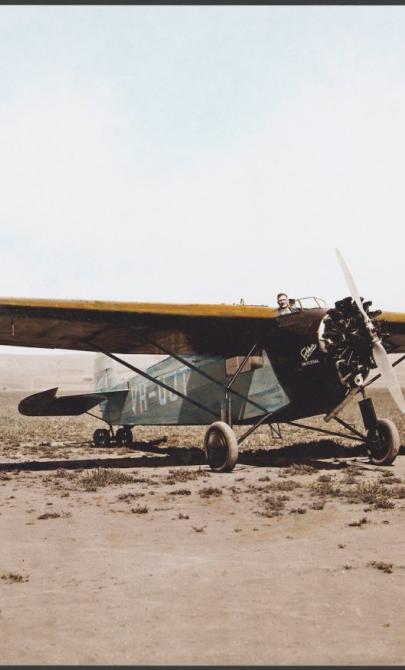 An old plane on the ground in a desert. A man is poking his head out from the cockpit and can be seen behind the wings of the plane