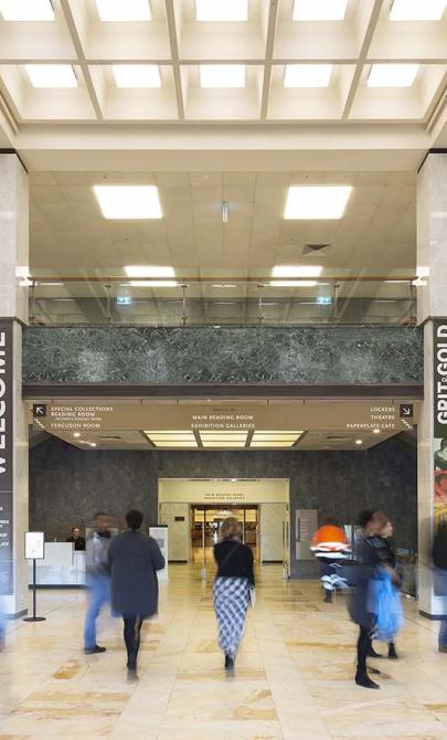 Large two-story room with cream-coloured marble floors, columns and stairs; black leather couches; lots of light from the ceiling and a balcony from the second floor looking over the room. On the columns are signs with directions and the word 'Welcome'. People are moving around the room.