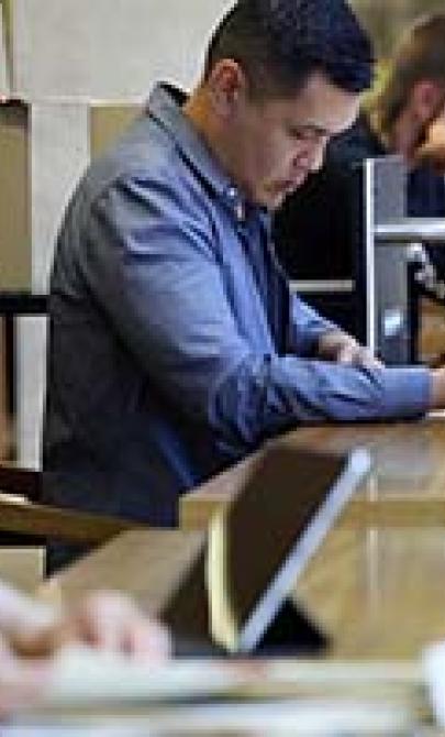 A group of people working in the special collections reading room. One person in the foreground is writing in a notebook, another in the middle is using a smartphone while taking notes, and others in the background are focused on their work.
