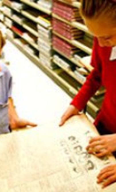 Three students in the library examining an old, oversized newspaper together. They are focused on the content, discussing and pointing out details. 