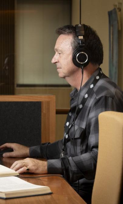 Man in a grey and black checkered shirt and headphones sitting at a desk, using a computer. To his left are two open books