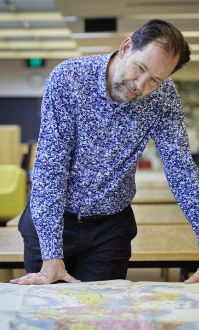 Man with brown hair and a grey and brown beard sitting at a table with a large map spread over it