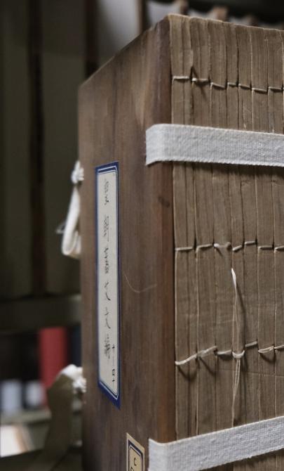 A large, old text with Chinese characters is bound with traditional thread stitching, sitting on a shelf, supported by a white fabric strap wrapped around its cover. The edges of the pages are uneven, showing wear with the stitching slightly frayed. Other colourful books with Chinese characters are lined up alongside it on the shelf.