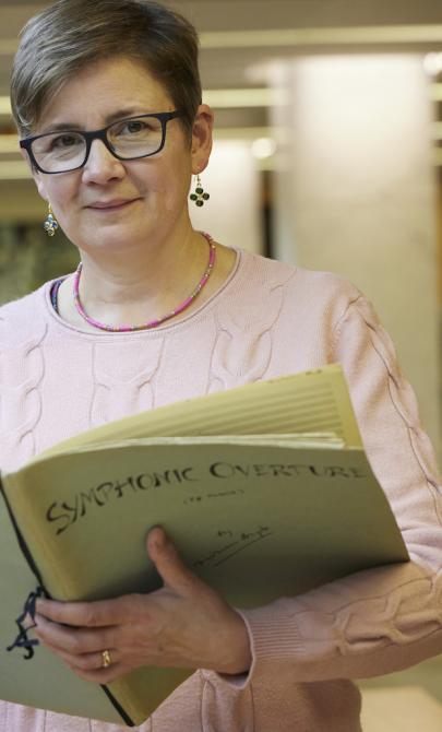 Celia Craig, a woman with very short brown hair, smiling and holding large book of sheet music with the title 'Symphonic Overture'