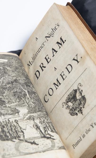 Book sitting open on a black cushion, showing an illustration of two armies having a stand-off in a clearing in a forest on the left page and on the right the title page for 'A Midsummer-Nights's Dream: 