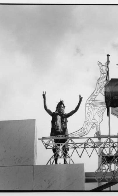 Two demonstrators stand on a structure at Parliament House, Canberra, raising their arms in the air beside an Australian Coat of Arms sculpture. A large peace flag is draped over the emblem. The black-and-white image captures the scene from below, with a cloudy sky in the background.
