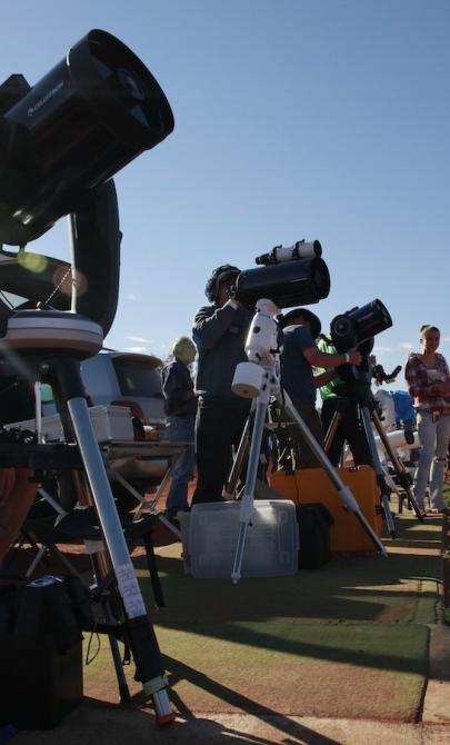 People in a red desert area sitting and standing behind large telescopes pointed towards the sky