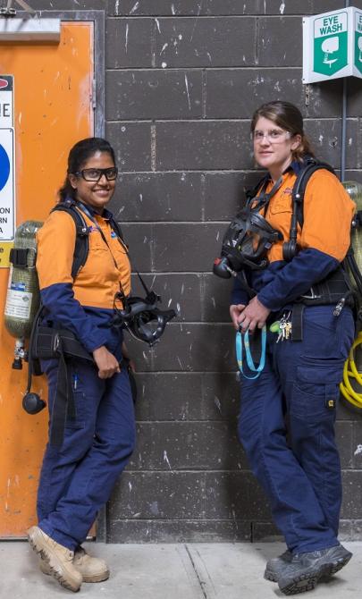Two women in navy and orange work wear with protective glasses, gas masks and tanks on their backs standing by a wall with an orange door and signs reading 'Danger Chlorine', 'Eye protection must be worn in this area', 'Liquid chlorine bulk storage' and more.