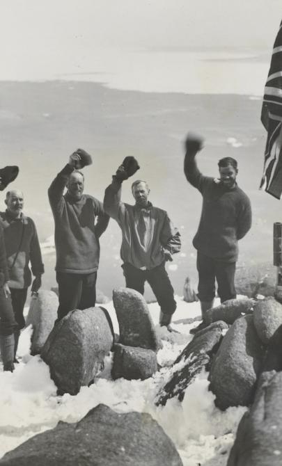 Group of explorers standing on the top of a snowing cliff overlooking a harbour, waving near a Union Jack flag