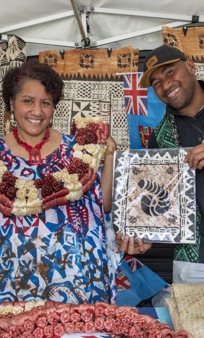 Man and woman running a stall selling Fijian products including woven mats, smiling at the camera