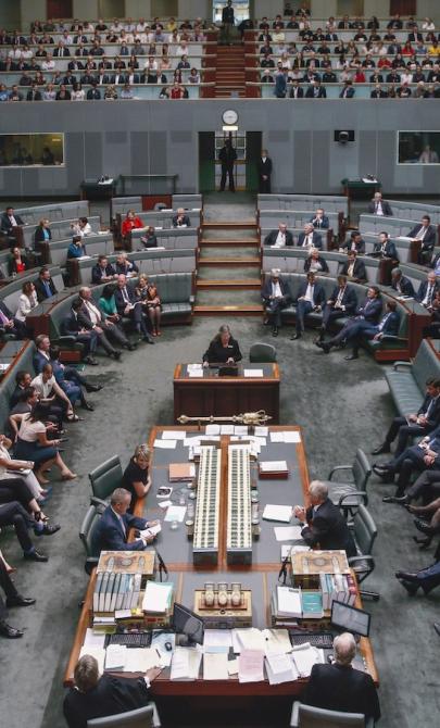 View of the Australian House of Representatives in Parliament House from slightly above. Members of the House of Representatives sit throughout the room, and members of the public sit in allocated areas