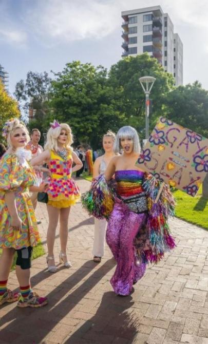 A group of four people who are very colourfully dressed, holding signs in a park. There are more groups of colourful people in the background.