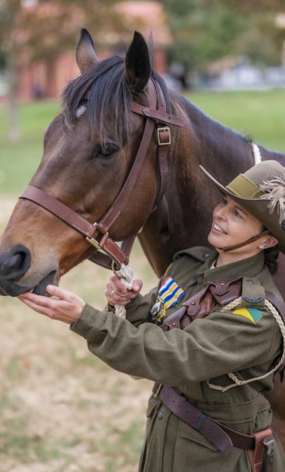 Female solider in uniform smiling and patting a military horse, with another soldier in uniform holding the reins of another horse nearby