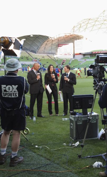 Three sports commentators with microphones standing on a soccer field in front of a large camera and small crew. 