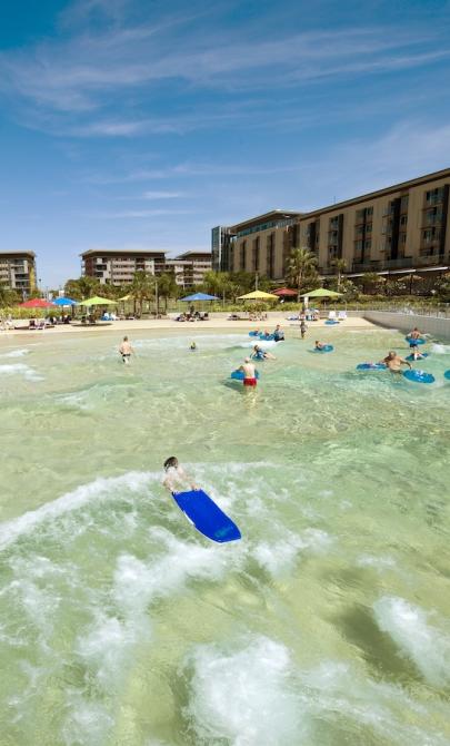 Adults and children playing in a large wave pool. Along the shore are dozens of large beach umbrellas with chairs underneath, and buildings surround the area