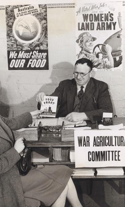 Man sitting behind a desk handing a booklet titled 'The Food Front' to a woman sitting on the other side of the desk. Signs around the pair and the desk read 'A Vital War Job - Join the Women's Land Army', 'We Must Share Our Food' and 'War Agricultural Committee'