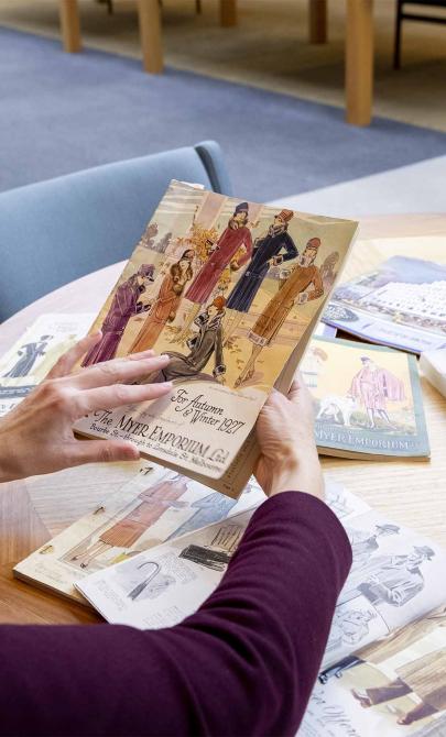 Looking over the shoulder of woman with black curly hair as she look at the cover of an old book about knitting and wool. More similar books are scattered across the table in front of her