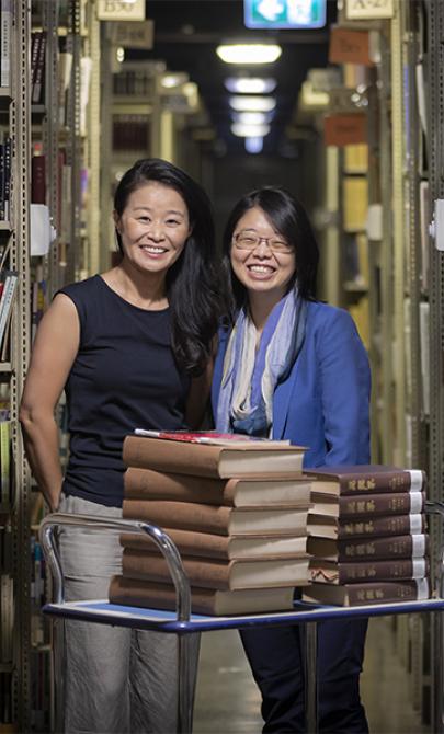 Two women standing behind a trolley with books on it in the Library stacks.