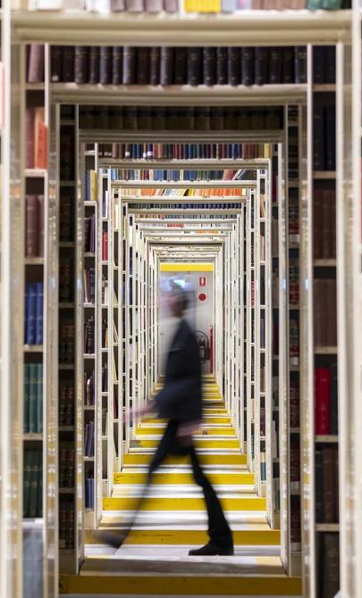 A blurred figure walking through a series of open shelving units at the Library. The shelves are lined with books, and the perspective creates a tunnel effect as the shelving repeats into the distance.